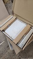 Metal cabinet pullout frame inside box with protective cardboard, showing assembly instructions beneath it.