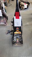 Front view of black Bissell ProHeat 2X Revolution carpet cleaner with red tape and lot tag attached on handle, placed on concrete floor.