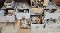 View of several plastic drawers and cardboard boxes filled with various euro cabinet hinges in different sizes and types, organized loosely.