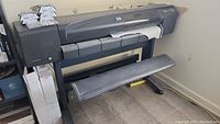 HP DesignJet 800 plotter printer with stand, several reams of plotter paper stacked beside it and boxes of ink cartridges placed on top of the plotter.