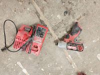 Photo showing Milwaukee red and black impact drill and red battery charger with power cord on a concrete floor.