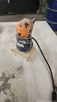 Ridgid palm router mounted on a wooden base with power cord wrapped around it, placed on a workbench.