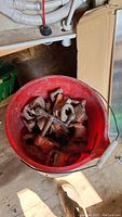 Red bucket containing multiple rusted pipe clamp ends with bolts and nuts in a workshop setting.