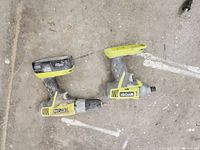 Photo of two Ryobi cordless power tools (a drill and impact driver) lying on concrete floor including two batteries, one attached to a tool and one detached.