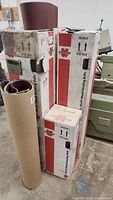 Four cardboard boxes labeled 3M Wood Sanding Belts stacked vertically beside a large roll of sandpaper