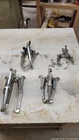 Four metal gear pullers arranged on a light-colored workbench surface. Varying sizes visible with typical three-arm configurations and central forcing screws.