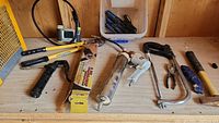 Tools on wooden surface including bolt cutter, grease gun, nail gun, hacksaw, screwdrivers, hammer, pliers, and handle grip.