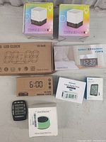 Full lot of eight clocks and timing devices in original factory packaging shown on a floor surface.