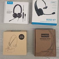 Four boxed communication headsets from different brands arranged flat: New Bee H360, Monoprice B350-XT, Mairdi BH-M97, and Mpow BH-M97.