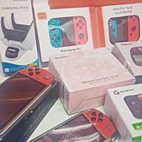 Photo showing various boxed gaming accessories including a charging dock, handgrips in red and blue, and a wireless controller for Nintendo Switch in white box.