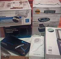 Photo showing multiple boxed home accessory items including garment steamer, wine tumblers, lint remover and lunch box