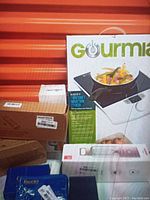 Photo showing boxed Gourmia 1800 watt induction cooker with packaging and additional smaller boxes and accessories around it.
