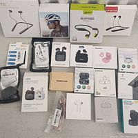 Top view of all 18 pieces including boxed sports headphones, wireless earbuds, and charging accessories in factory packaging laid out on a flat surface.