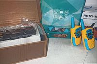 Two yellow and blue walkie talkies standing upright next to box with geometric pattern, additional boxed items in background.