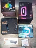 Photo showing the six items in their original packaging arranged on the floor, including the disco ball light double speaker, Oracolo Roller, FM110 speaker, wireless RGB speaker, XSound GO, and the small round Bluetooth speaker/CD player.