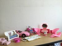 Barbie pink Corvette, pink motorcycle, white loveseat with cushions, small pink coffee table, vanity mirror, two modern pink doll chairs, and bedding pieces displayed on tabletop.