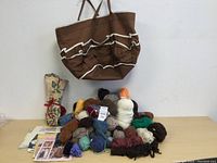 Large brown knitting tote bag hung on wall above pile of assorted yarn skeins and rolled needle case on table.
