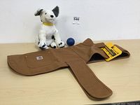Large brown Carhartt firm duck insulated dog chore coat laid flat with a white stuffed dog toy and a blue ball toy placed on a table.