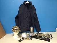 Full view showing black men's water resistant jacket hanging, camo Realtree Max 5 hat on stand, Duck Dynasty drawstring backpack flat, fishing gear, tumbler, and sunglasses arranged below jacket.