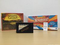Photo showing the Settlers of Catan box, Scrabble box, and Dominoes box arranged in a row on a table.
