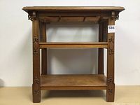 Front view of the carved hardwood table showing carved floral details on the legs and two lower wooden shelves.