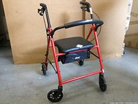 Red metal walker with cushioned black seat, ergonomic black handles, and four wheels.