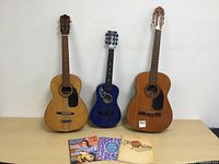 Front view of three acoustic guitars, two natural wood finish, one small blue, all standing upright against white background with guitar books in front