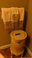 Yellow cylindrical accent table with a drawer on top and a cabinet door below, floral painted decoration, placed near corner of room with two beige decorative towels hanging above.