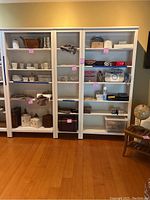Three white wooden bookcases side by side, filled with various household items (not included in sale). Each has six shelves and a paneled side design.