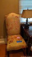 Full view of the floral upholstered chair with wooden legs and the table lamp on a side table next to it.