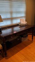 Full view of dark wooden desk with three drawers placed under a window with blinds, lamp on the side, and some small objects on the desktop (not included).