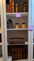White shelf containing multiple faux book boxes resembling antique books, two jar candles, and decorative metal candle holder.