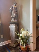 Concrete female figurine statue on top of tall concrete pillar, floral arrangement with white flowers and green leaves in a clay pot with saucer placed beside them. Whole lot shown in corner against wall.