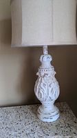 Full view of one lamp showing distressed white ornate carved base and beige fabric lampshade