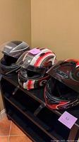 Three motorcycle helmets lined up on a black shelf showing their front and side profiles, with protective visors attached, varying colors, and visible scratches.