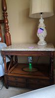 Full view of the wooden side table with white marble top, lamp on top, turned legs, and a power plug visible behind it.