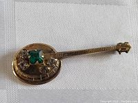 Front view of gold toned banjo brooch pin with green square rhinestone and clear rhinestones on the banjo body.