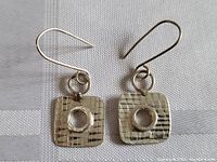 Pair of sterling silver dangle earrings with textured square pendants and central circular cutout, shown on fabric background.