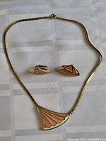 Photo showing the vintage Trifari peach enamel pendant necklace with matching clip earrings laid flat on a white cloth. Pendant has an art deco fan design with peach and cream enamel on gold-tone trim. The earrings are triangular clip-ons with matching enamel and trim.