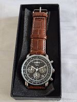 Full view of watch in black presentation box showing brown leather strap and dial