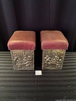 Front view of two antique brass fireplace fender seats with reddish leather seats and embossed brass panels depicting figures in historic dress