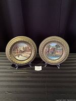Front view of two round handpainted plaster wall plaques with antique brass frames showing detailed colorful outdoor scenes.