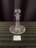 Clear glass decanter with stopper on top, shown on striped cloth background.