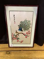 Full view of framed embroidery artwork featuring peacocks on a flowering branch, with Asian characters, set against a light background.