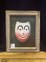 Full view of the framed oil painting of a clown face mask on a dark background with signature visible.