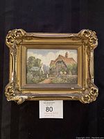 Framed oil painting showing a cottage and garden scene with ornate gold frame, displayed on black background.
