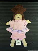 Full front view of the Cabbage Patch doll lying flat, showing pink knitted dress and pants with duck embroidery and brown yarn hair.