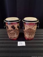 Front view of pair of conga drums showing detailed artwork of huts, palm leaves, and shorelines on tan and red painted surfaces with black tuning hardware.