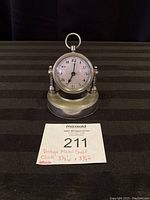 Front view of clock showing round face with Arabic numerals and metal base.