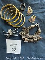 View of entire jewelry lot on dark blue fabric including six gold-tone bangles, two brooches (dragonfly and spider), pair of flame-shaped earrings, and charm bracelet with animal figures.